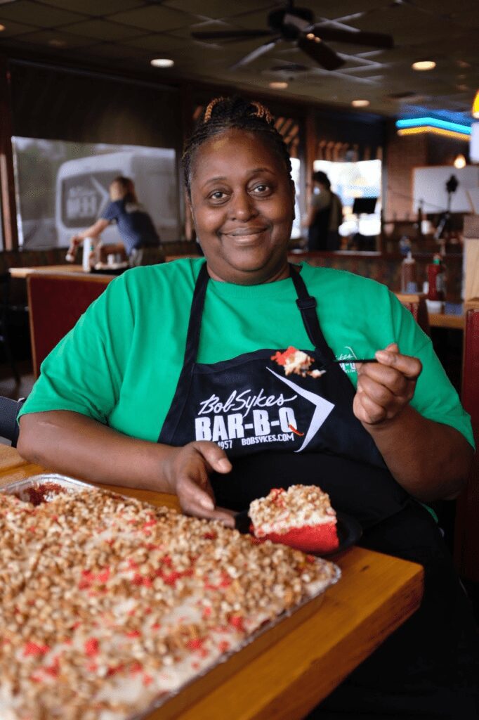 Woman wearing a black apron and green shirt eating cake